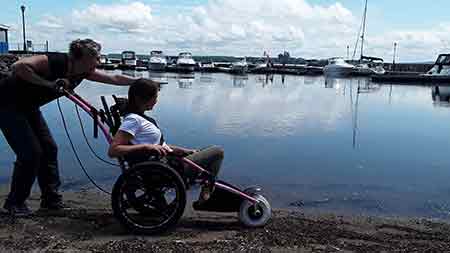 A person pushing a person in a hippocampe on sand on the Pictou Waterfront with boats in background.